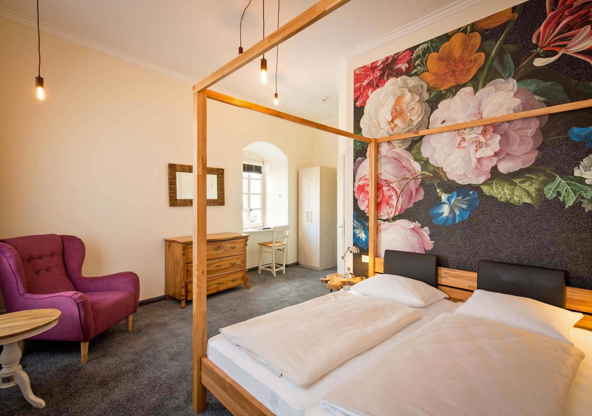 Ein helles, beige gestaltetes Doppelzimmer im Deutschherrenhof. Der dunkle Teppichboden und helle Holzmöbel bilden einen harmonischen Kontrast. Im Raum stehen ein Himmelbett und ein lilafarbener Sessel. Hinter dem Bett befindet sich eine schwarze Wand mit einer Fototapete bunter Blumen.