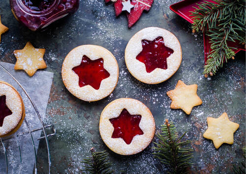 Drei Spitzbuben-Plätzchen liegen auf einem weihnachtlich dekorierten Untergrund. Im Hintergrund befinden sich weitere Plätzchen auf einem Abkühlrost, Tannenzweige ragen ins Bild, und ein Glas mit Marmelade steht daneben.