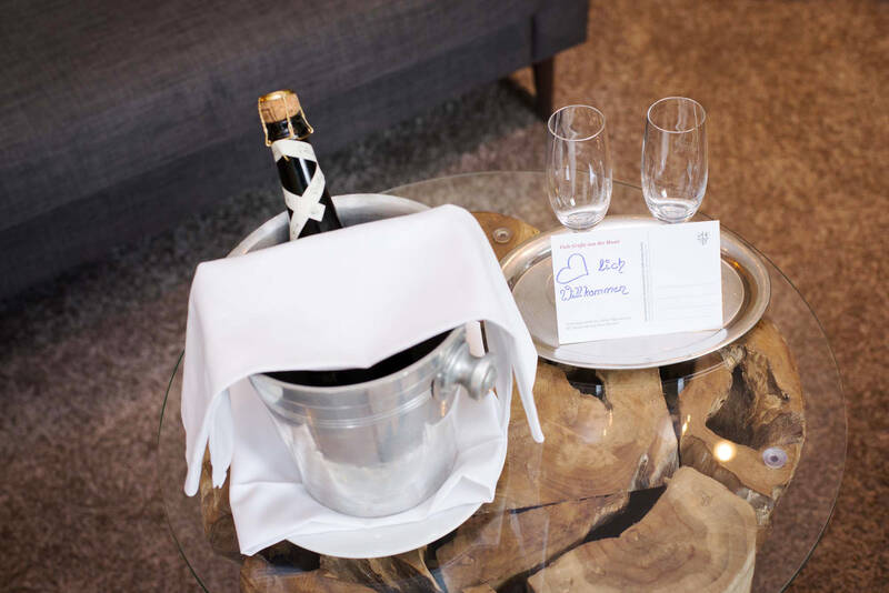 Auf dem Beistelltisch neben einem Sofa steht eine Flasche Sekt in einem abgedeckten Sektkühler. Daneben liegt ein Tablett mit zwei leeren Sektgläsern und einer Postkarte, auf der ‚Herzlich Willkommen‘ steht.