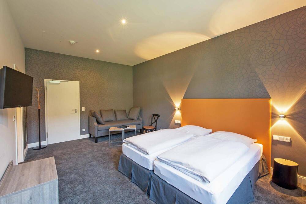 Ein hell beleuchtetes Zimmer im Deutschherrenhof. Die Wand hinter der Eingangstür und dem Bett ist grau-grün gemustert. Neben der Tür stehen ein Sofa und ein kleiner Beistelltisch. Das Doppelbett hat ein knallorangefarbenes Kopfteil. Leuchten über den Nachttischen spenden gemütliches Licht.