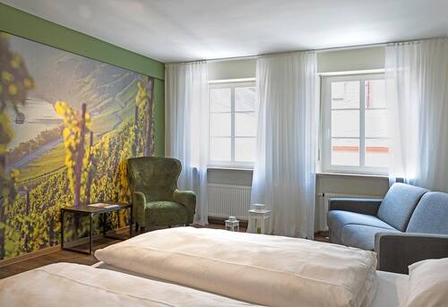 In einem hell erleuchteten Zimmer zeigt eine Fototapete eine Landschaft mit Weinbergen und der Moselschleife. Vor der Tapete steht ein grüner Sessel mit einem kleinen Beistelltisch. Neben dem Sessel gibt es ein graues Sofa und ein Doppelbett im Raum.