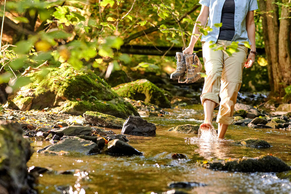 Eine Frau steht mit den Füßen in einem Bach. Sie hat die Hose hochgekrempelt und trägt ihre Wanderschuhe in der Hand. Mit dem rechten Fuß spritzt sie Wasser in die Luft.