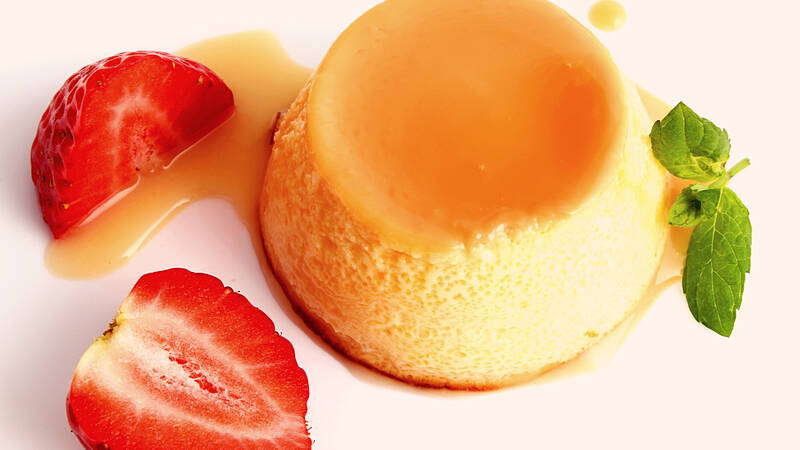 Zwei Erdbeerscheiben liegen neben einem Stück Panna Cotta auf einem Teller. Das Dessert ist mit zwei frischen Minzblättern dekoriert.