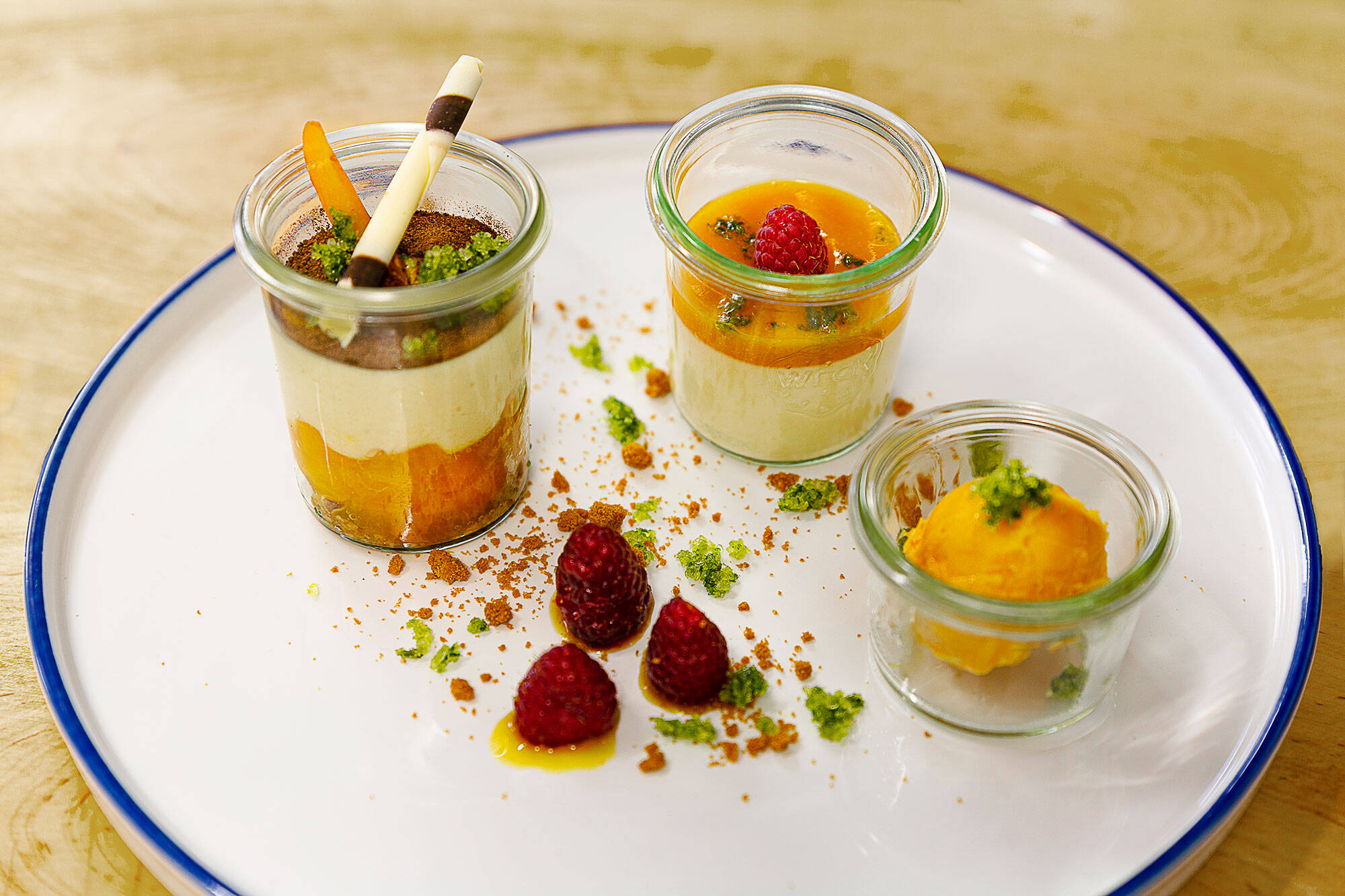 Drei kleine Gläser mit verschiedenen Desserts stehen auf einem Teller. Der Teller ist mit Gewürzen und Himbeeren dekoriert.