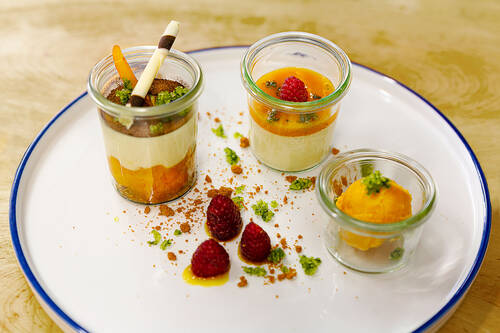 Drei kleine Gläser mit verschiedenen Desserts stehen auf einem Teller. Der Teller ist mit Gewürzen und Himbeeren dekoriert.