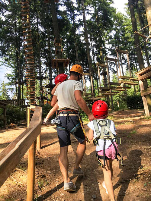 Ein Mann mit Kletterausrüstung und Helm geht durch ein Waldstück. Auf dem Arm trägt er ein kleines Mädchen, ebenfalls mit Ausrüstung und Helm. An seiner Hand läuft ein etwas älteres Mädchen, auch in Kletterausrüstung. Sie gehen gemeinsam in Richtung Kletterparcours.