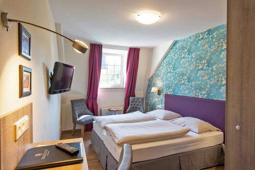 Blick in ein farbenfrohes Doppelzimmer im Deutschherrenhof. Ein Doppelbett mit lila Kopfteil steht vor einer türkis gemusterten Blumentapete. Am Fenster hängen bodenlange lilafarbene Vorhänge. Davor steht eine Sitzecke mit zwei gemütlichen Stühlen.