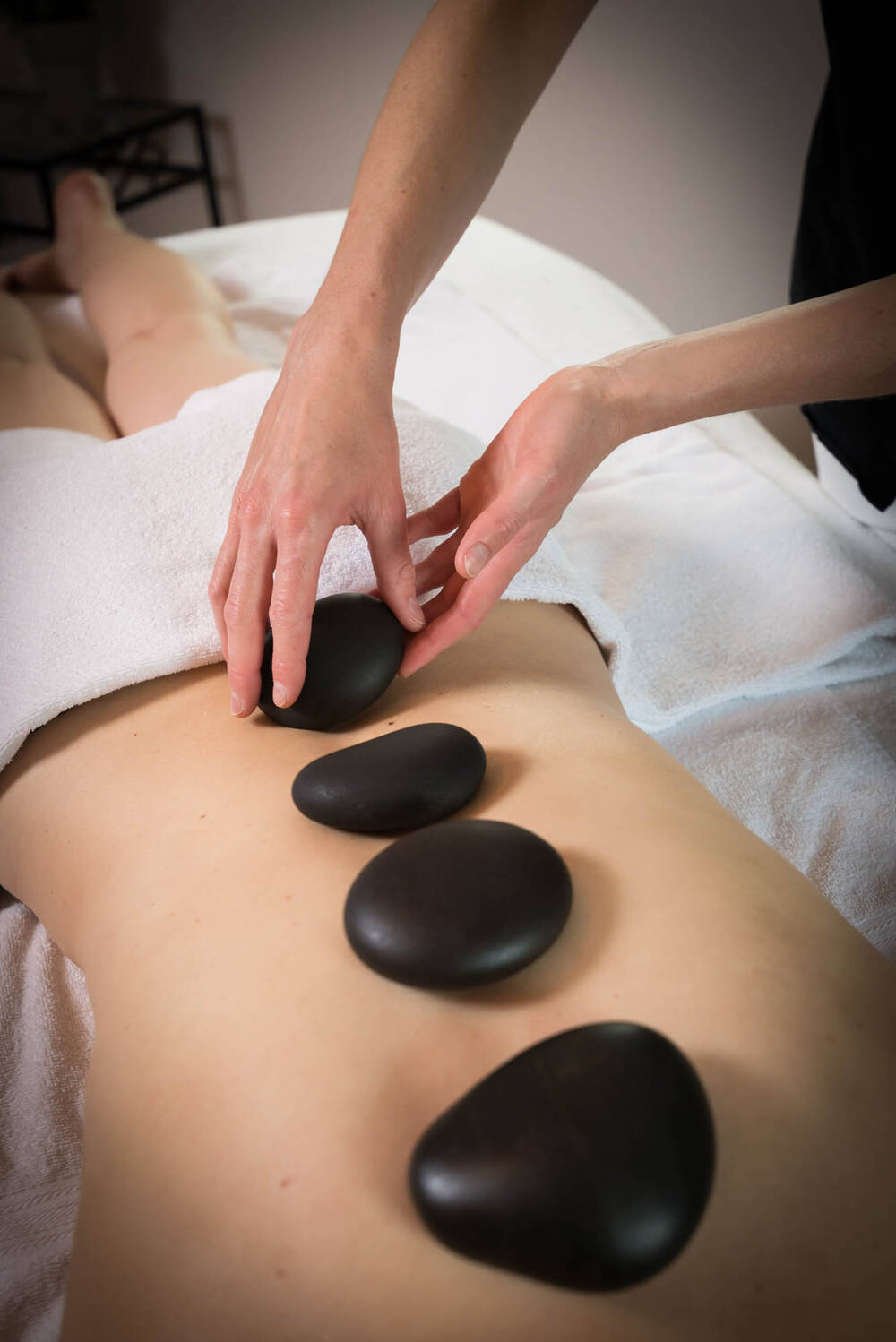 Eine Frau liegt mit einem Handtuch um die Hüfte auf einer Massageliege und erhält eine Hot-Stone-Massage von einer Mitarbeiterin.