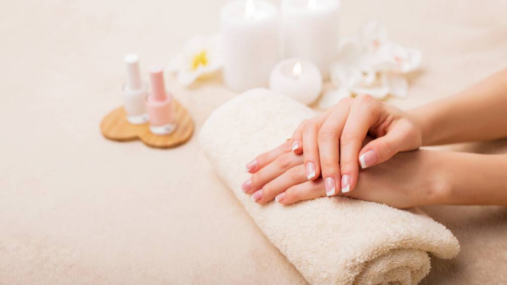 Zwei gepflegte Hände liegen gefaltet auf einem gerollten Handtuch. Die Nägel sind frisch lackiert. Im Hintergrund sind Kerzen, Nagellacke und Blüten von Blumen zu sehen.