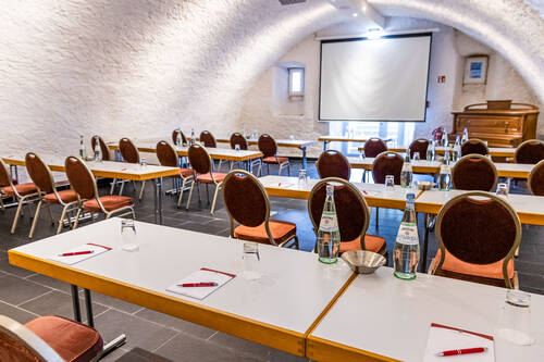 Im Gewölbekeller des Deutschherrenhofs stehen Tische und Stühle in Reihen hintereinander, alle ausgerichtet auf eine vorne angebrachte Leinwand. Auf den Tischen liegen Notizblöcke mit Kugelschreibern sowie leere Gläser und jeweils zwei Flaschen Sprudel.