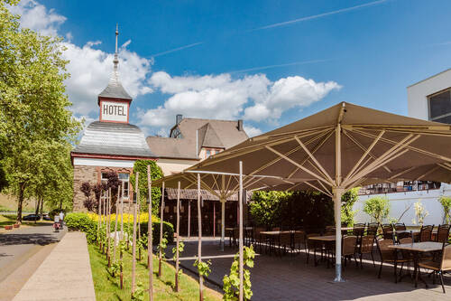Die Sonnenterrasse des Hotels Deutschherrenhof: An der Mauer entlang der Straße wachsen einige Weinreben. Dahinter stehen große Sonnenschirme, die den gemütlichen Sitzgruppen darunter angenehmen Schatten spenden.