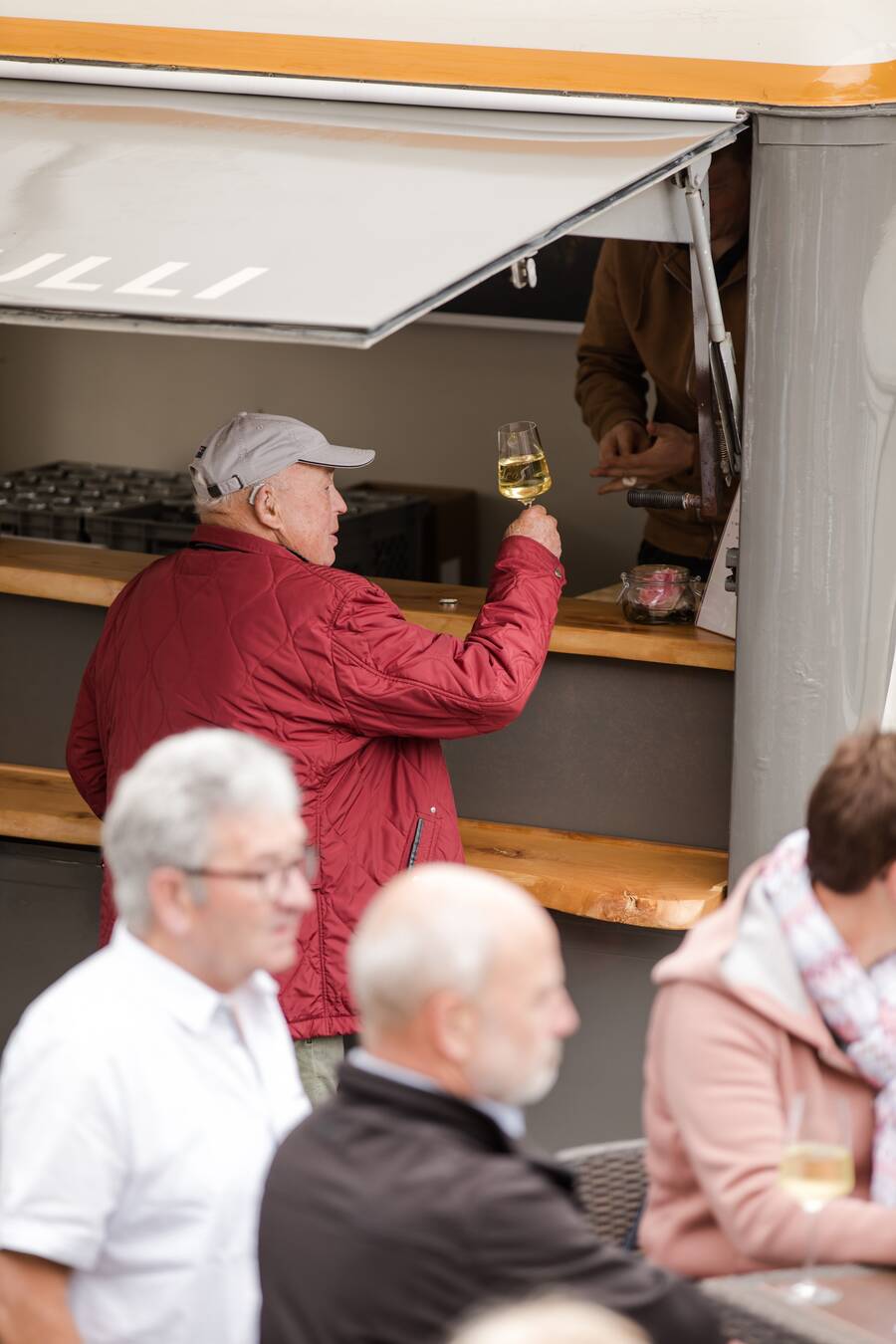 Ein älterer Mann in roter Jacke hält ein gefülltes Weinglas in der Hand. Er steht vor einem Wein-Bulli und stößt freundlich mit dem Winzer an.