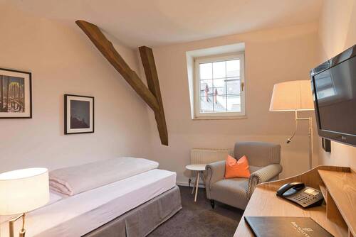 „Weiß gestrichenes Einzelzimmer im Deutschherrenhof mit einem grauen Sessel und einem Einzelbett. Über dem Bett hängen zwei Fotos. Auf dem Sessel liegt ein oranges Kissen als farblicher Akzent. In der Ecke des Raums ragen alte Holzbalken aus der Wand.