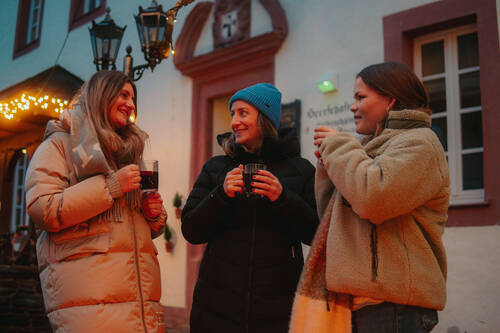 Drei Frauen stehen im Innenhof des Hotels, eingepackt in Winterkleidung. Jede hält eine dampfende Tasse Glühwein in der Hand und lächelt sich gegenseitig an.