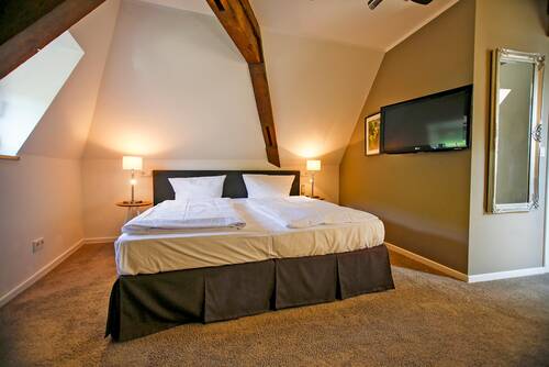 Ein Doppelbett mit weißer Bettwäsche steht in einem grün-weiß gestrichenen Hotelzimmer im Deutschherrenhof. Sichtbare Holzbalken ragen aus der Decke. Der Raum ist mit Teppichboden ausgelegt. An der Wand hängen ein Spiegel und ein Fernseher. Nachttischlampen spenden warmes, gemütliches Licht.