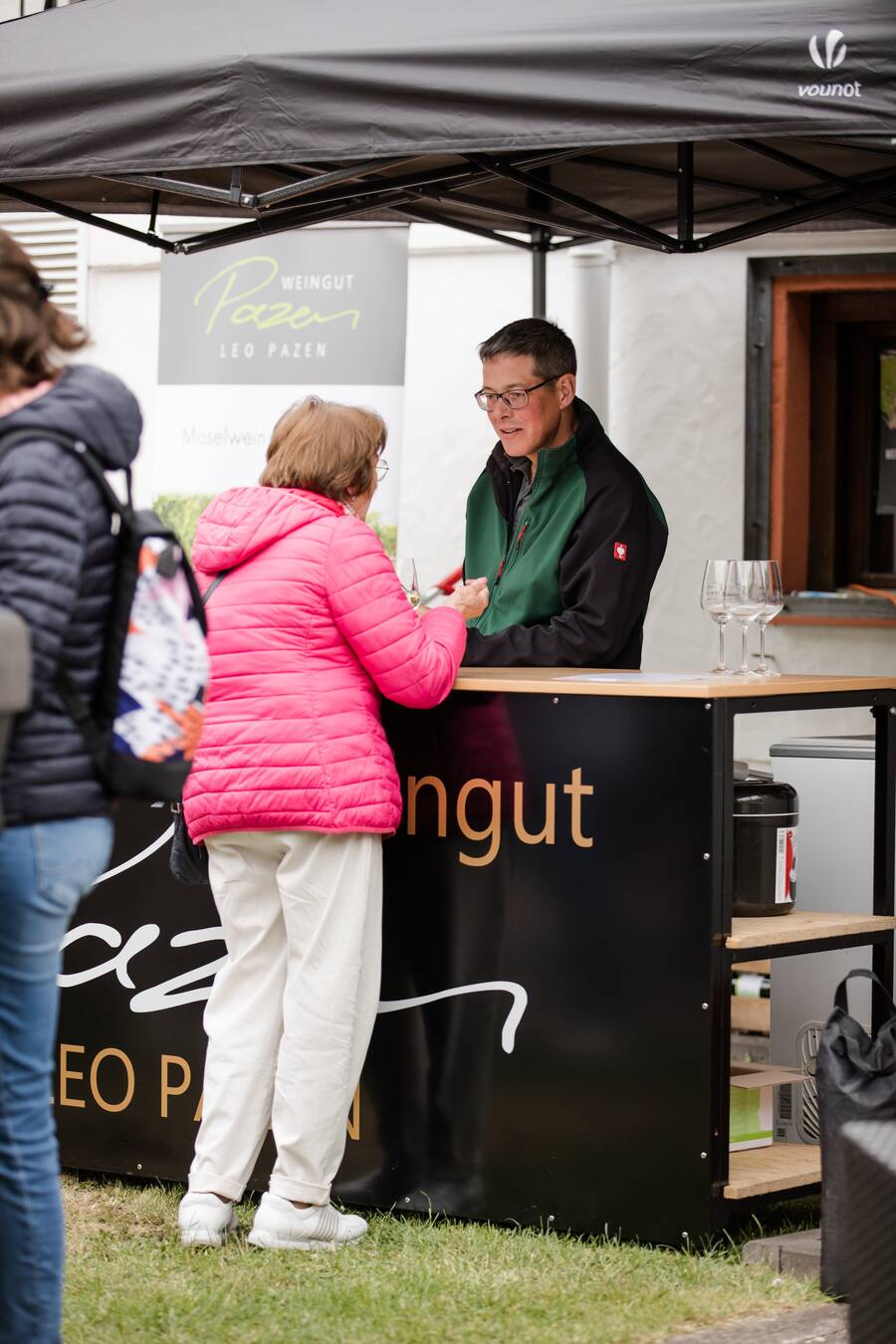 Am Verkaufsstand des Weinguts Leo Pazen steht der Winzer hinter der Theke und schenkt einer Frau in einer pinken Steppjacke Wein ein.