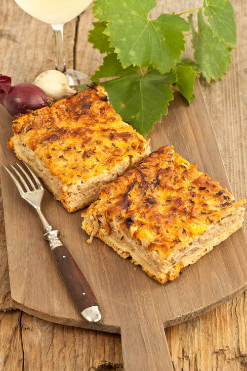 Zwei Stücke Zwiebelkuchen liegen auf einem Holzbrett, daneben eine Gabel. Im Hintergrund sind der Stiel eines Weinglases, Zwiebeln und Weinblätter zu sehen.