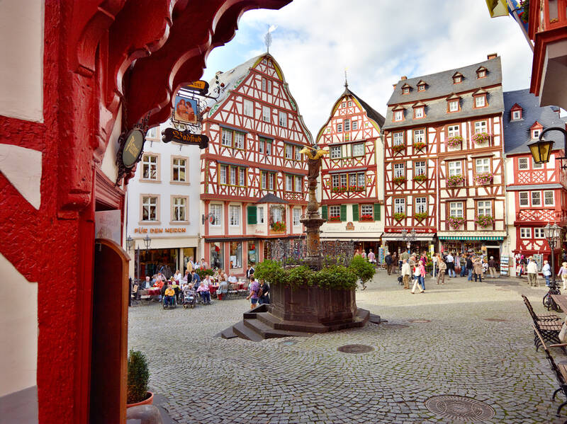 Auf dem Marktplatz von Bernkastel stehen historische Fachwerkhäuser rund um einen Brunnen. Zahlreiche Touristen bewegen sich über den Platz.