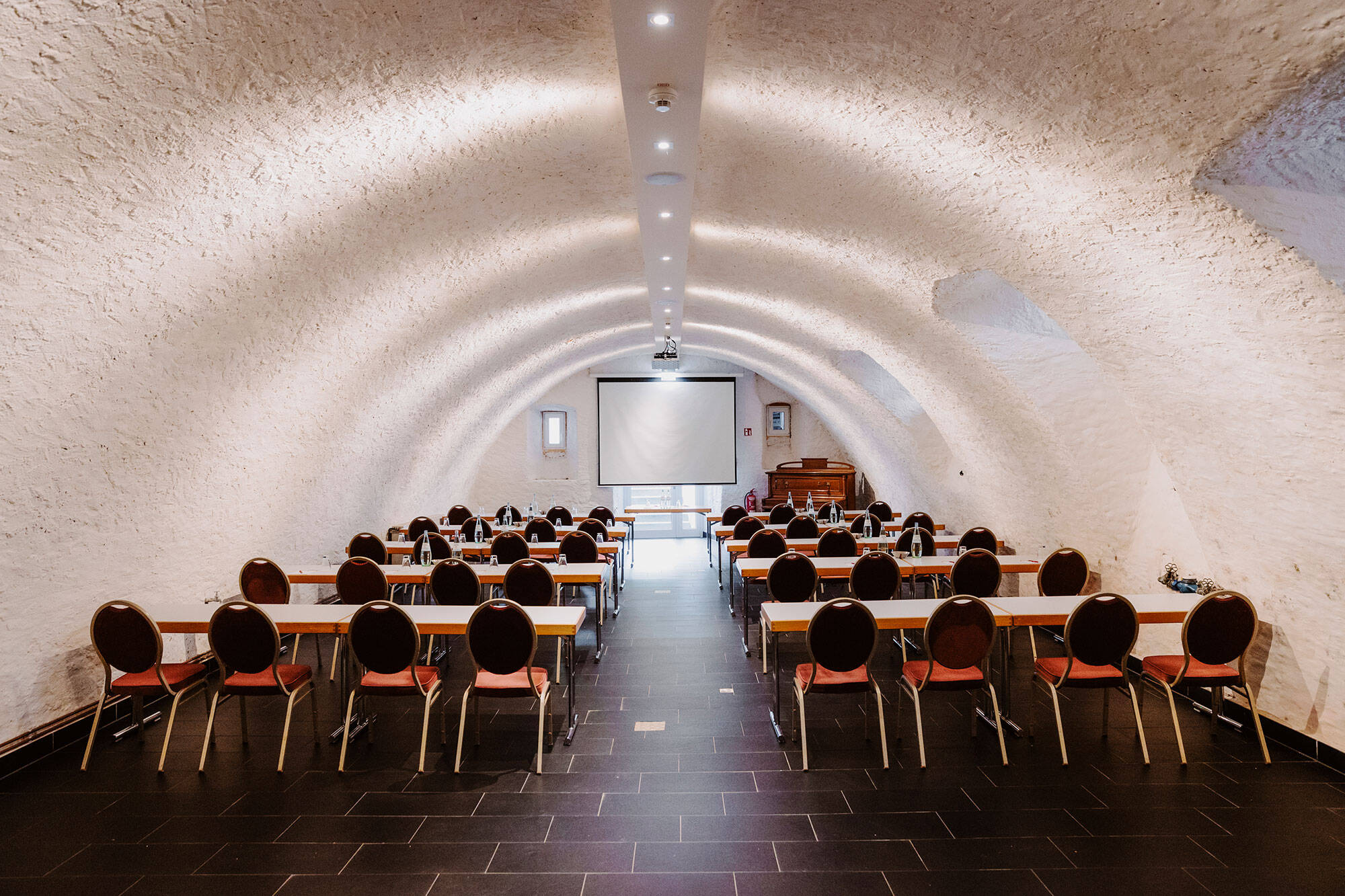 Im Gewölbekeller des Deutschherrenhofs stehen Stühle und Tische in Reihen hintereinander. Sie sind alle auf eine vorne angebrachte Leinwand ausgerichtet.