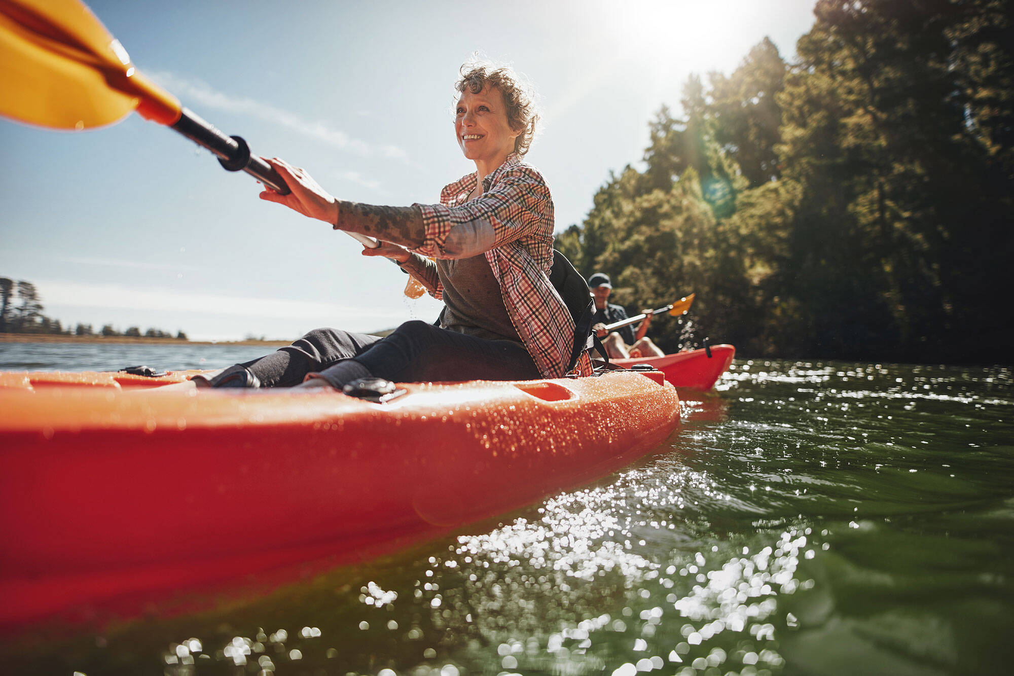Eine Frau sitzt in einem roten Kanu und paddelt mit einem Lächeln über das Wasser. Im Hintergrund scheint die Sonne.