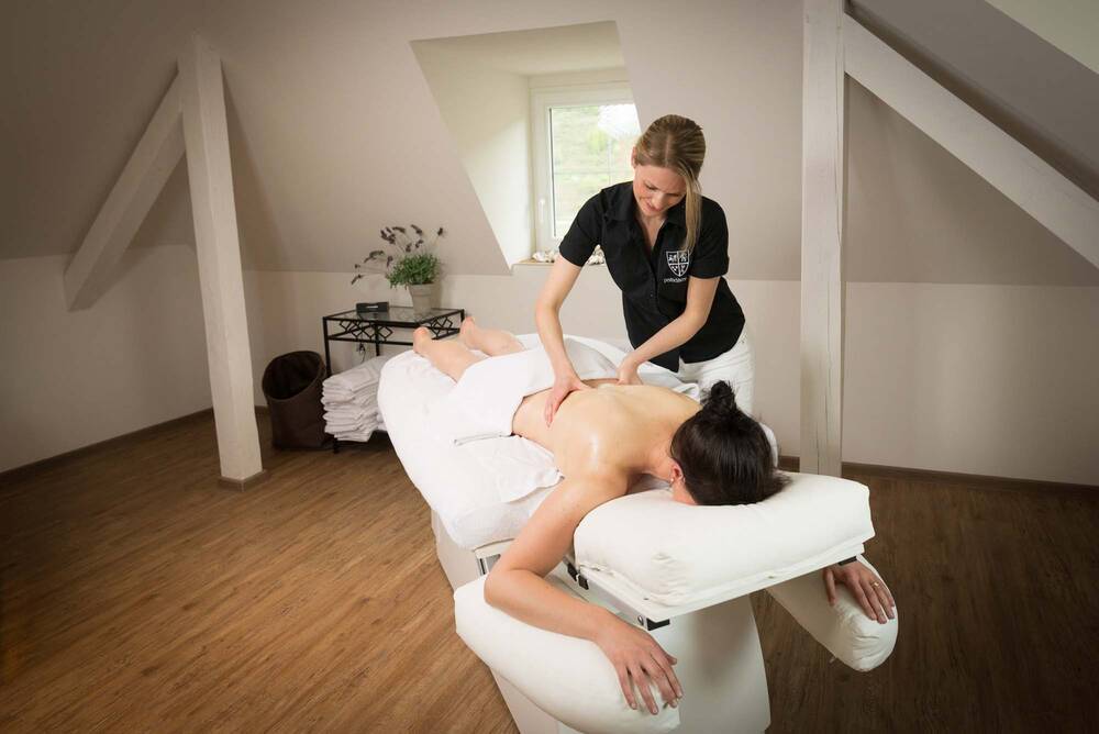 Eine Frau liegt mit einem Handtuch um die Hüfte auf einer Massageliege und erhält eine Hot-Stone-Massage von einer Mitarbeiterin.