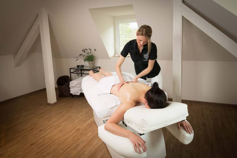 Eine Frau liegt mit einem Handtuch um die Hüfte auf einer Massageliege und erhält eine Hot-Stone-Massage von einer Mitarbeiterin.