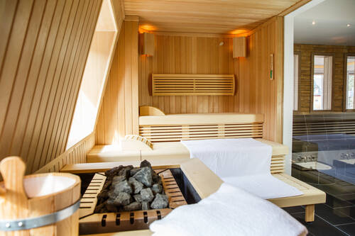 Blick in die Sauna des Deutschherrenhofs. Auf den drei Holzstufen liegen große weiße Handtücher. Im vorderen Bildrand sind der Holzkübel und die Saunasteine für den Aufguss zu sehen. Durch die Glaswand und Fenster wird die Sauna von natürlichem Licht erleuchtet.
