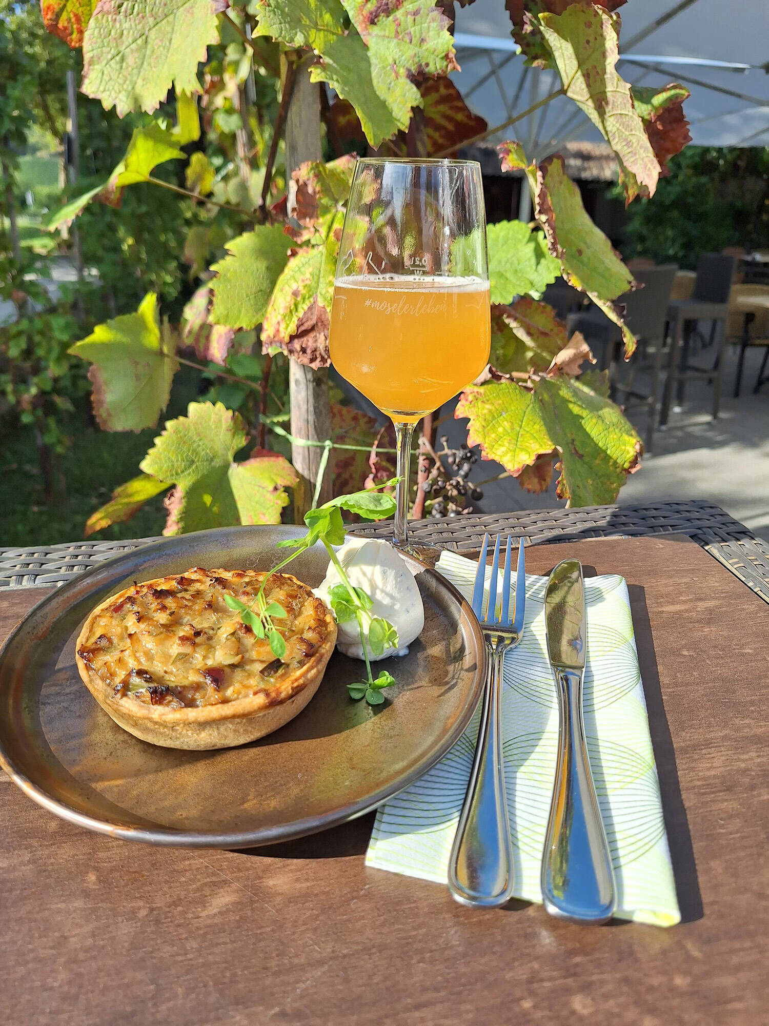 Ein Stück Zwiebelkuchen liegt auf einem Teller auf dem Tisch. Daneben liegen Besteck und eine Serviette, ein Glas mit frischem Federweißer steht davor. Im Hintergrund sind eine Weinrebe und die Terrasse des Hotels zu sehen.