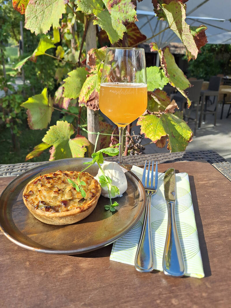 Ein Stück Zwiebelkuchen liegt auf einem Teller auf dem Tisch. Daneben liegen Besteck und eine Serviette, ein Glas mit frischem Federweißer steht davor. Im Hintergrund sind eine Weinrebe und die Terrasse des Hotels zu sehen.
