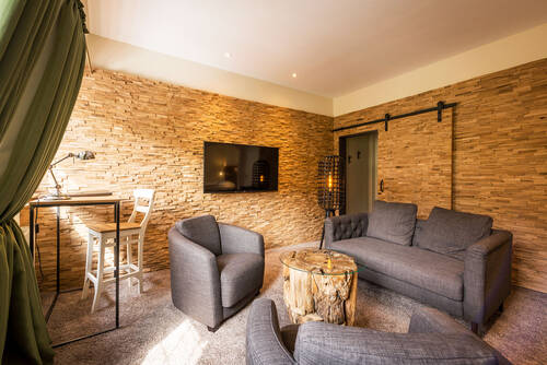 Wohnzimmer der Suite im Deutschherrenhof. Zwei graue Sessel und ein graues Sofa stehen um einen kleinen Beistelltisch. An der Wand hängt ein Fernseher. Rechts im Bild befindet sich die Tür zu einem Schrank.