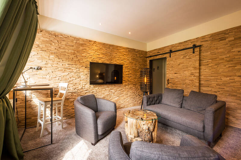 Wohnzimmer der Suite im Deutschherrenhof. Zwei graue Sessel und ein graues Sofa stehen um einen kleinen Beistelltisch. An der Wand hängt ein Fernseher. Rechts im Bild befindet sich die Tür zu einem Schrank.