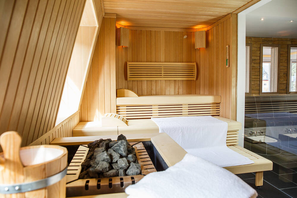 Blick in die Sauna des Deutschherrenhofs. Auf den drei Holzstufen liegen große weiße Handtücher. Im vorderen Bildrand sind der Holzkübel und die Saunasteine für den Aufguss zu sehen. Durch die Glaswand und Fenster wird die Sauna von natürlichem Licht erleuchtet.
