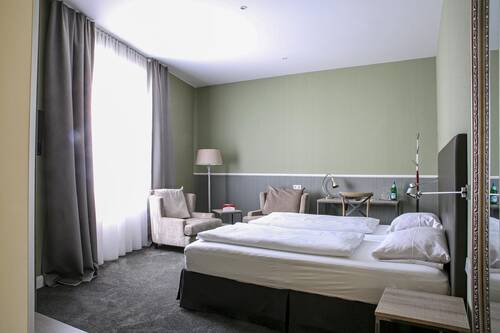 Grün-braun gestaltetes Rieslingzimmer im Deutschherrenhof. Neben dem Doppelbett befindet sich eine Sitzecke mit zwei Sesseln, einer Lampe und einem kleinen Tisch. Bodentiefe Vorhänge am Fenster lassen warmes Tageslicht ins Zimmer.