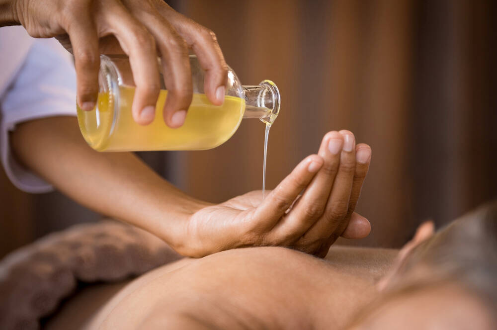 Eine Person liegt auf dem Bauch. Eine andere Person gießt Massageöl aus einer Glasflasche in ihre Handfläche, um es aufzutragen.