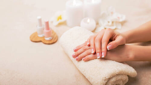 Zwei gepflegte Hände liegen gefaltet auf einem gerollten Handtuch. Die Nägel sind frisch lackiert. Im Hintergrund sind Kerzen, Nagellacke und Blüten von Blumen zu sehen.