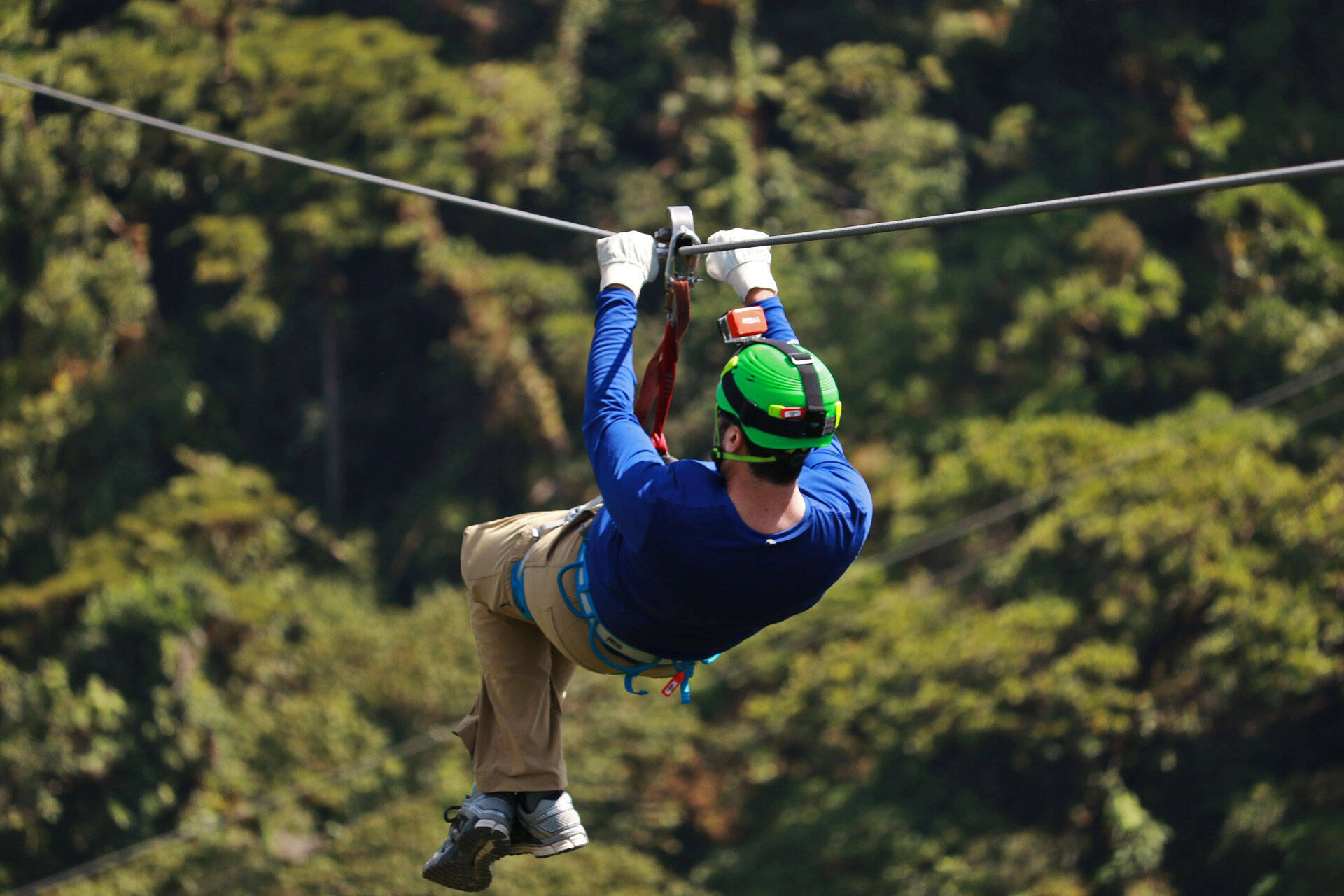 Ein Mann mit Kletterausrüstung und Helm, an dem eine Actionkamera befestigt ist, rutscht an einer Seilrutsche über einen Wald.