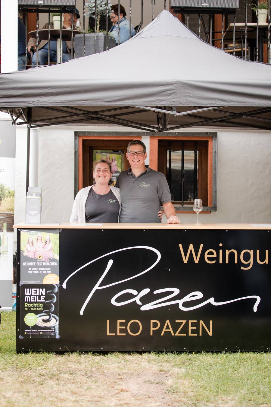 Das Ehepaar Pazen steht hinter einer Theke mit der Aufschrift ‚Weingut Leo Pazen‘. Sie umarmen sich und lächeln in die Kamera. Der Verkaufsstand befindet sich im Innenhof des Hotels Deutschherrenhof.
