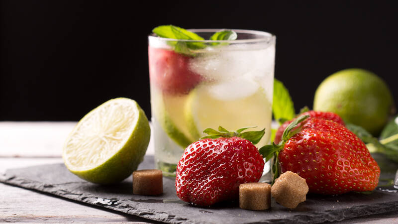 Auf einer Schieferplatte liegen Limetten, Erdbeeren und Würfel aus braunem Zucker dekorativ arrangiert. Dahinter steht ein Glas, gefüllt mit Eiswürfeln, Erdbeeren, Limetten, Zitronenscheiben und einem erfrischenden Drink.