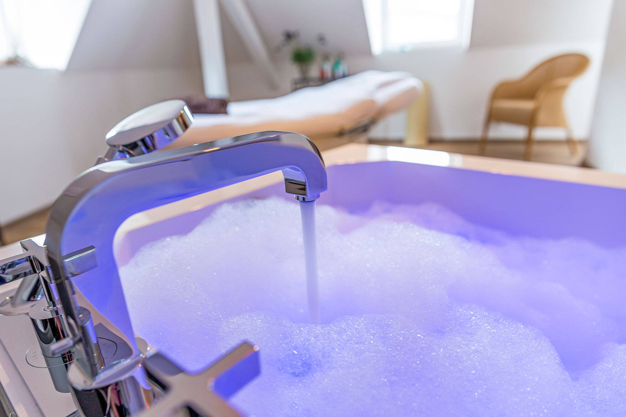 Eine Badewanne wird mit Schaumwasser aufgefüllt. Der Wasserhahn ist im Vordergrund des Bildes sichtbar, während im Hintergrund eine Kosmetik- oder Massageliege zu sehen ist. Das Schaumwasser in der Wanne wird von lilafarbenem Licht erleuchtet.