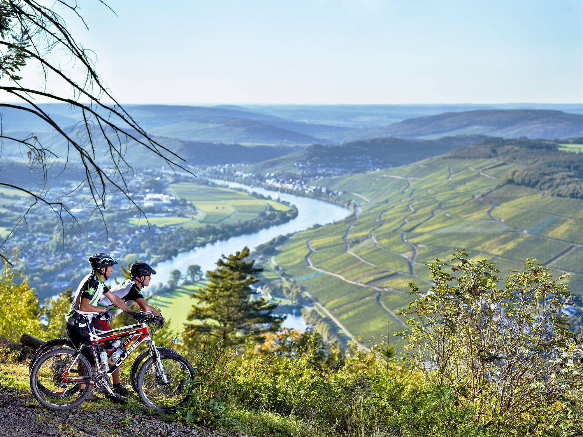 Zwei sportlich gekleidete Radfahrer stehen mit ihren Mountainbikes an einem Aussichtspunkt und blicken auf das wunderschöne Moselpanorama.