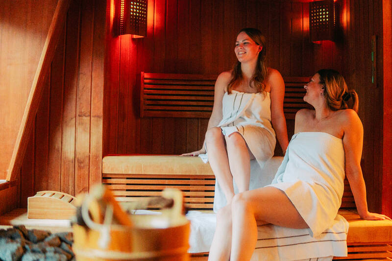 Zwei Frauen, in weiße Handtücher gewickelt, sitzen in der Sauna und lächeln.