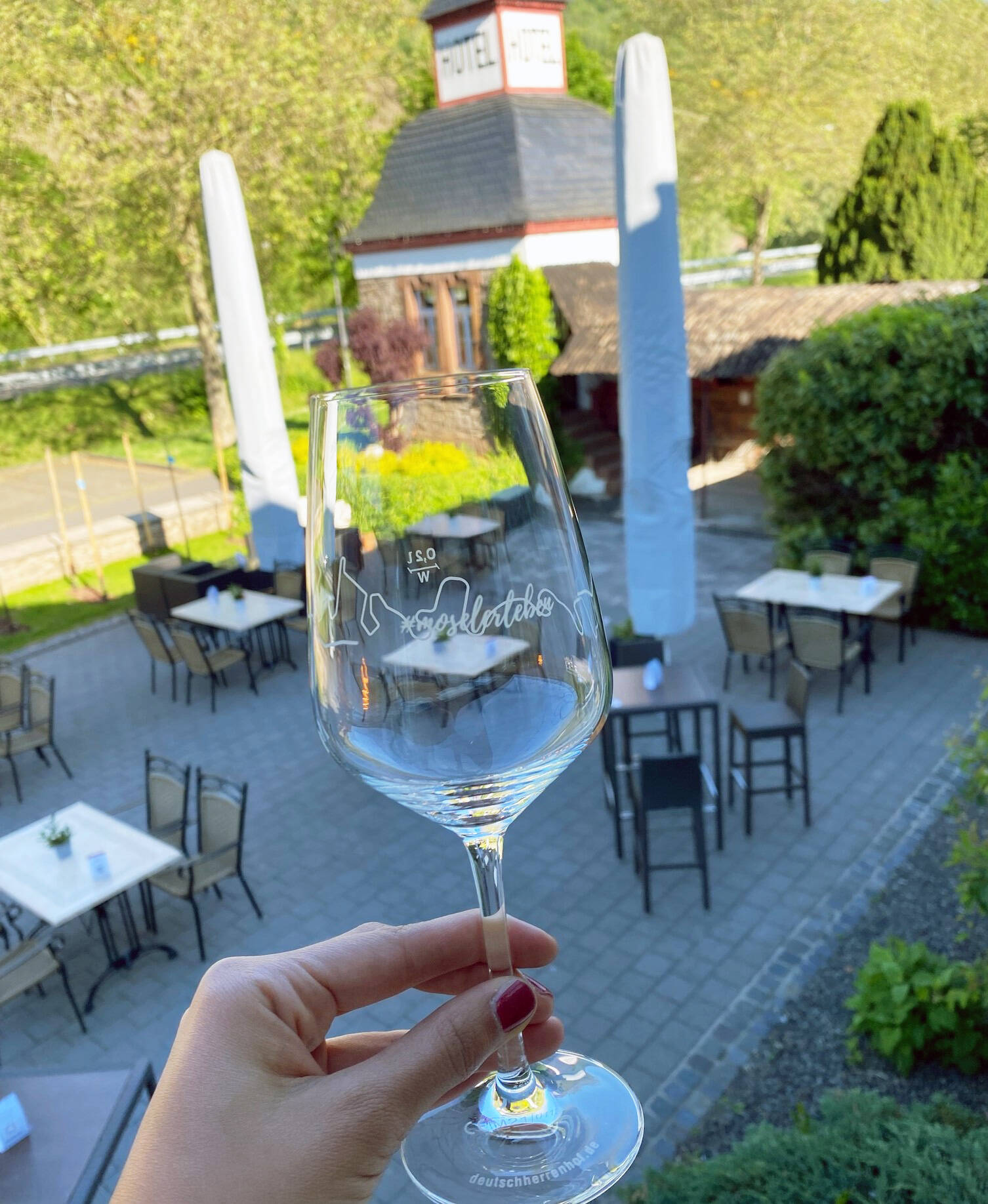 Eine Hand hält ein Weinglas mit dem Aufdruck "Mosel erleben" des Deutschherrenhof. Im Hintergrund ist die Terrasse des Deutschherrenhof zu sehen.