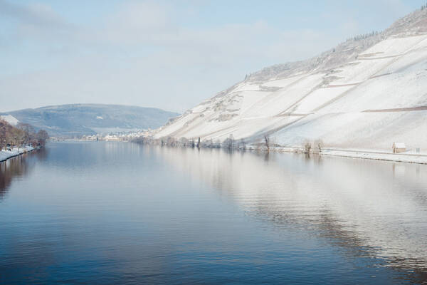 Die schneebedeckten Weinberge spiegeln sich in der ruhigen Mosel. Der Himmel ist strahlend blau.