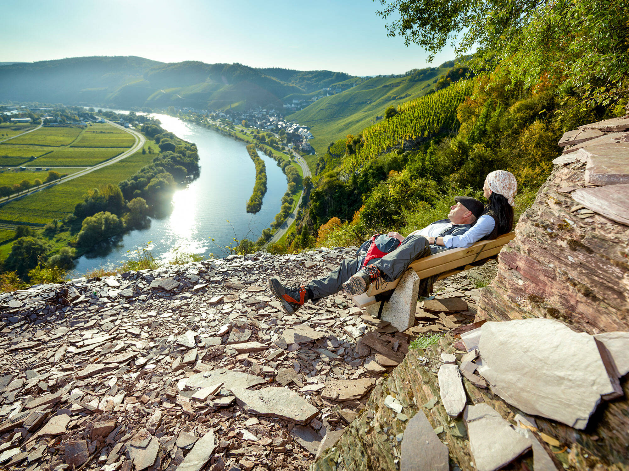 Eine Frau sitzt auf einer Bank auf einem Berg. Ihr Partner liegt mit dem Kopf in ihrem Schoß. Seine Beine ragen über den Rand der Bank. Im Hintergrund ist die Moselschleife zu sehen. Das Wasser ist ruhig, der Himmel blau, die Sonne scheint.