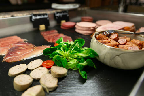 Auf Schieferplatten ist eine vielfältige Auswahl an Wurstsorten arrangiert. In einer weiteren Schüssel sind kleine Stücke Rohesser zu sehen. Zur Dekoration sind auf den Platten einige Blätter Feldsalat und ein paar Tomatenscheiben verteilt.